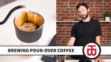 Warren grinds coffee beans, places a filter inside the pour over coffee dripper, and places it onto a tumbler. Hot water is poured around the outside edges first. Warren stops pouring once the entire surface is no longer dry. Warren resumes pouring when the surface appears to stop moving. He keeps the coffee saturated and foamy, pouring a little more every few seconds, until the tumbler is full. He tastes the coffee, demonstrates how to clean the dripper, and shows it on top of the French Press.